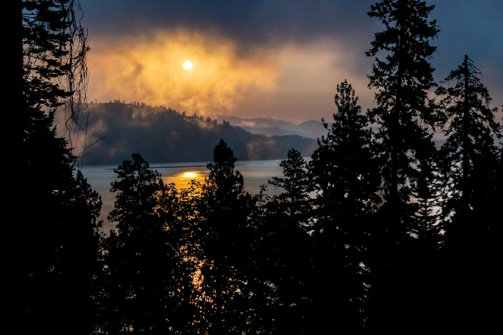Photograph; A morning sun orange with wildfire smoke shines beneath dark rain clouds above a mountain lake, in the foreground silhouetted pine trees.