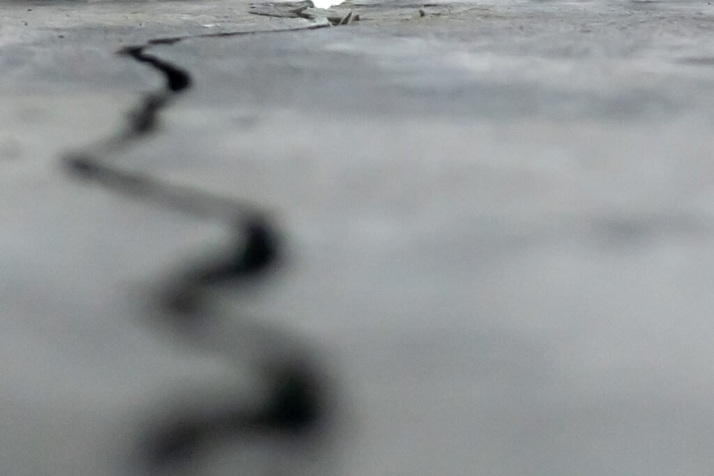 Photograph; a crack in a dark gray cement wall leading up to a chip revealing the sky.