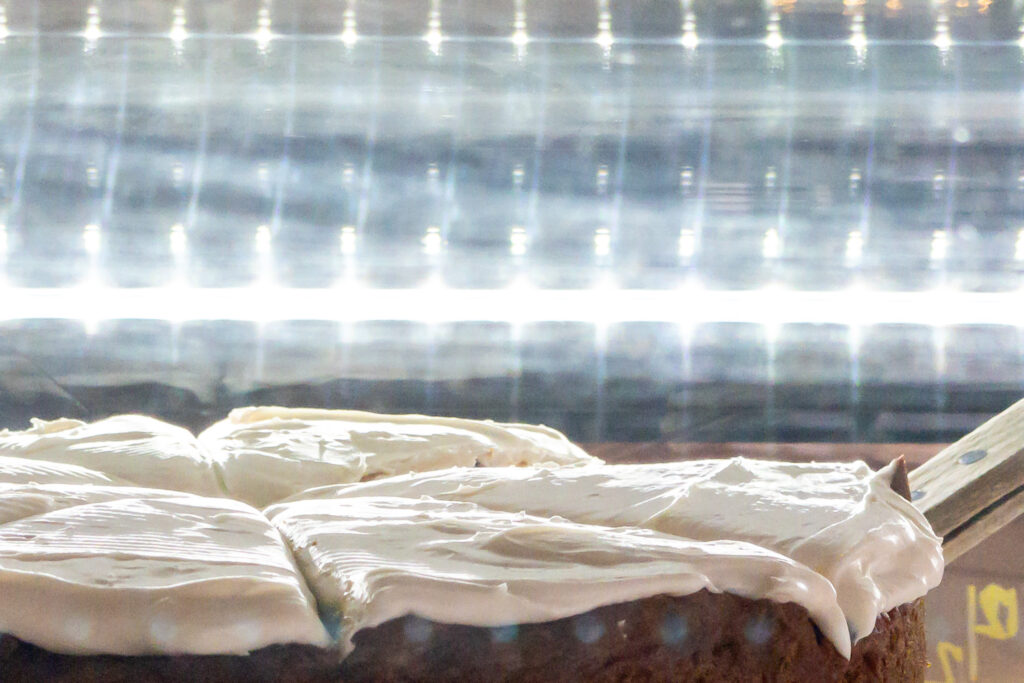 Photograph; a frosted pumpkin spice cake under a string of lights in a cafe display case.