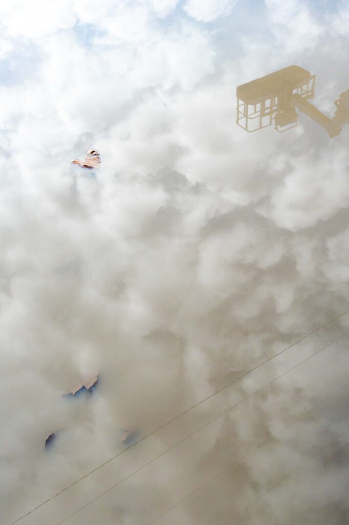 Photograph; A reflection of sky and clouds, a bucket truck bucket, and power lines in a puddle of mud, leaves, and a rock.