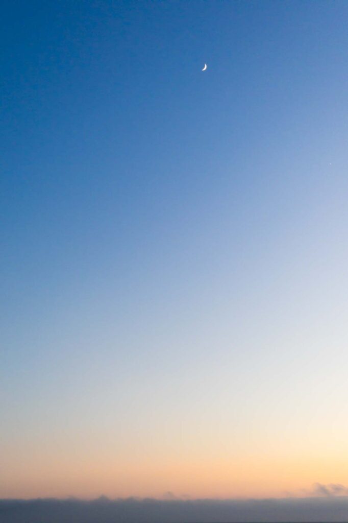 Photograph; A crescent moon high above a layer of low clouds at sunset.