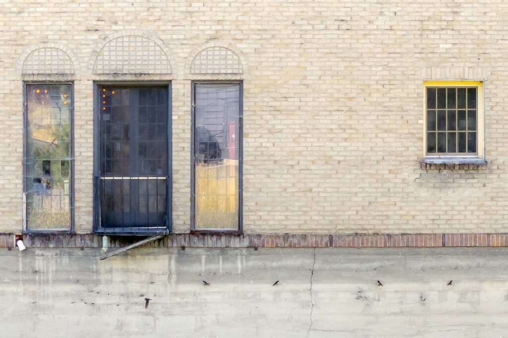 Photograph; a brick wall, three windows and a double door, below four swallows hang on a cement wall, one flying by.