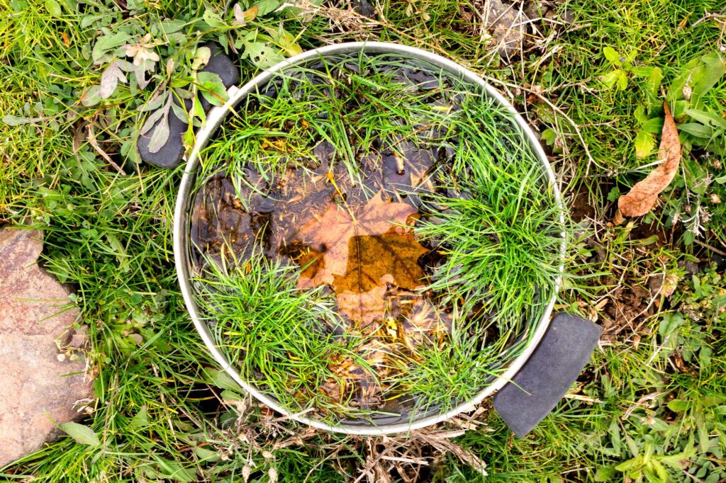 Photograph; A soup pot left outdoors sitting next to a stone, full of dirt, grasses, and autumn leaves with only its rim and handles above the earth.