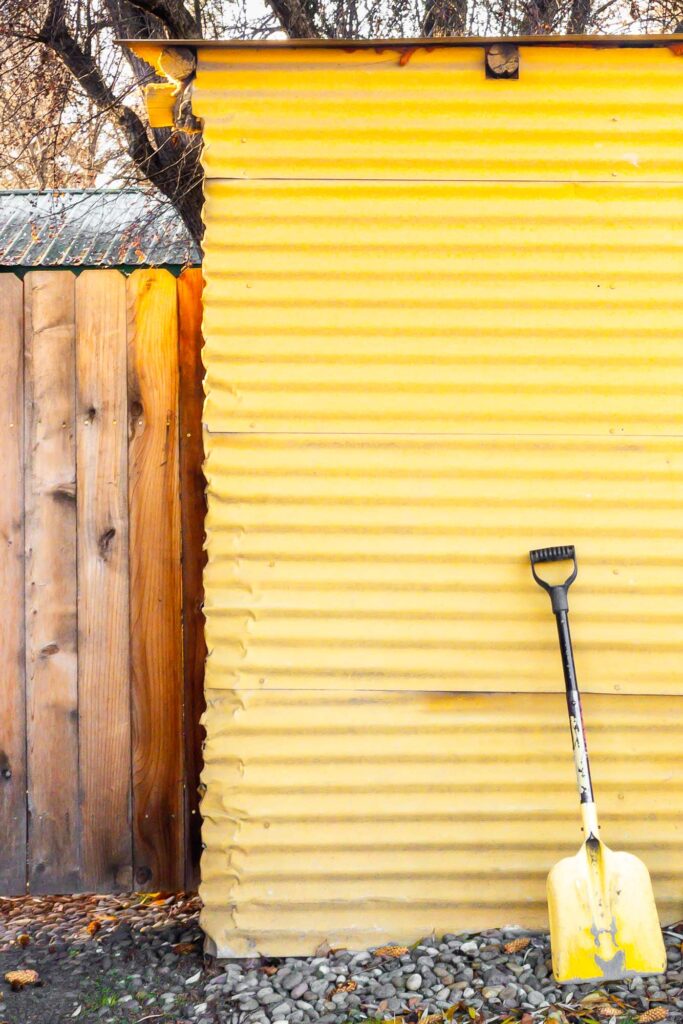 Photograph; A yellow shovel with a black handle, propped against a yellow shed standing in river rock next to closed gate and the roof of a house.