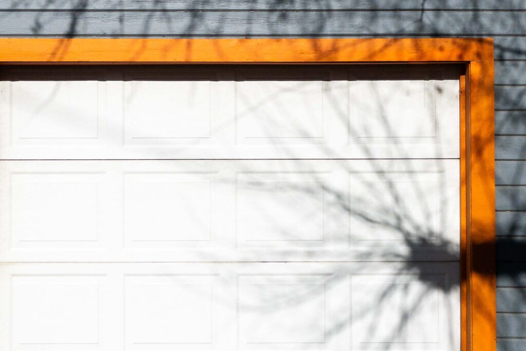 Photograph; A white garage door with orange trim, and a blue gray wall with shadows of bare tree branches surrounding the door and on the door on the right.