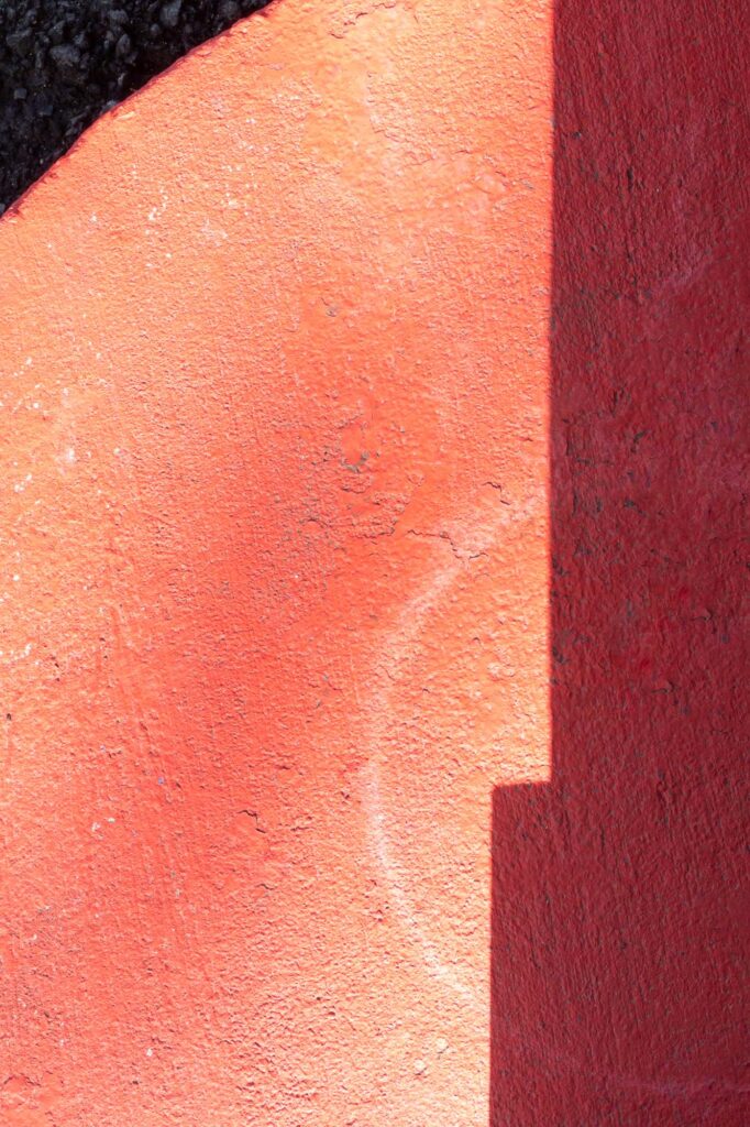 Photograph of a curb with fading red paint with a water ring from a pot. A building shadow is on the right and in the upper left the curve of the curb and beyond the curve gravel.