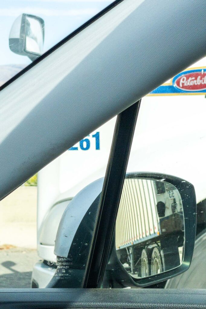 Photograph from the inside of a car looking out the front and passenger window being passed on the right by a semi truck with a reflection of the semi in the review mirror.