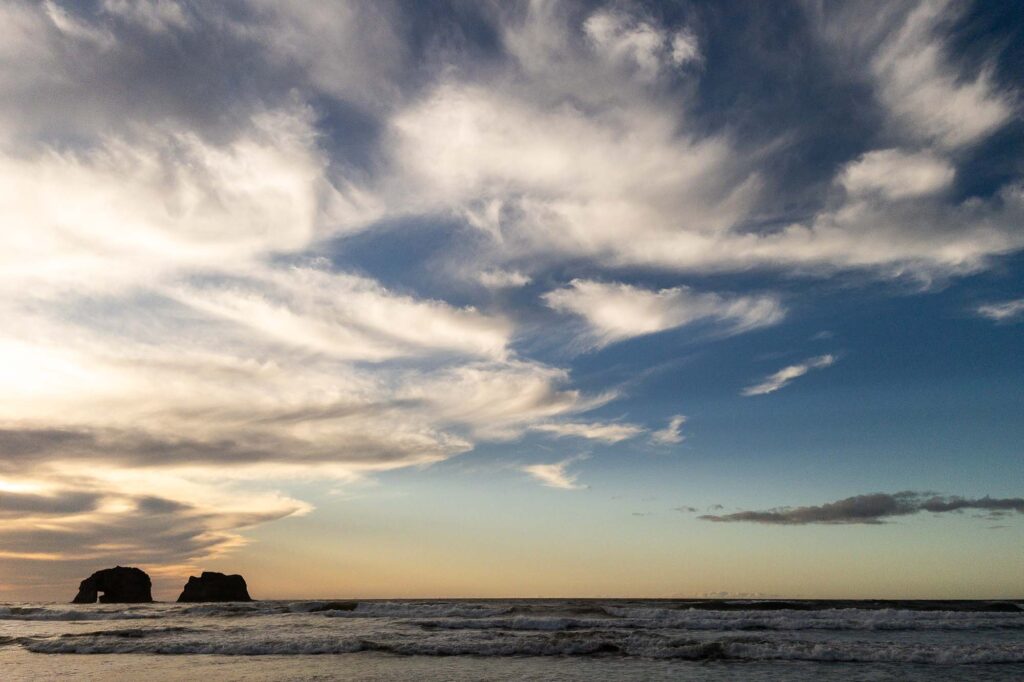 Photograph of wispy clouds from the beach at Sunset over the ocean waves and two large rocks, one with an arch, on the left out in the ocean.