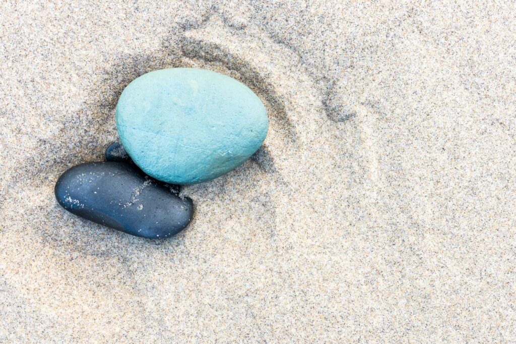 Photograph of three stones one large that is green, one medium that is black, and one small that is black, deposited on the beach in a depression in the sand from the outgoing tide.