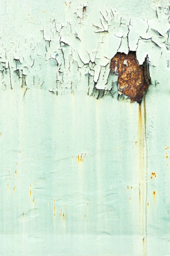 photograph of the side of a railway car with faded green paint. Across the top the paint is peeling, with a rusty hole dripping leaving a streak of rust running down the side.