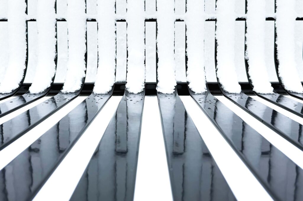 Photograph of a bench with metal slats  taken from above, the seat slats covered in snow, the bench back slats reflecting the snow covered seat slats. With snow on the ground.