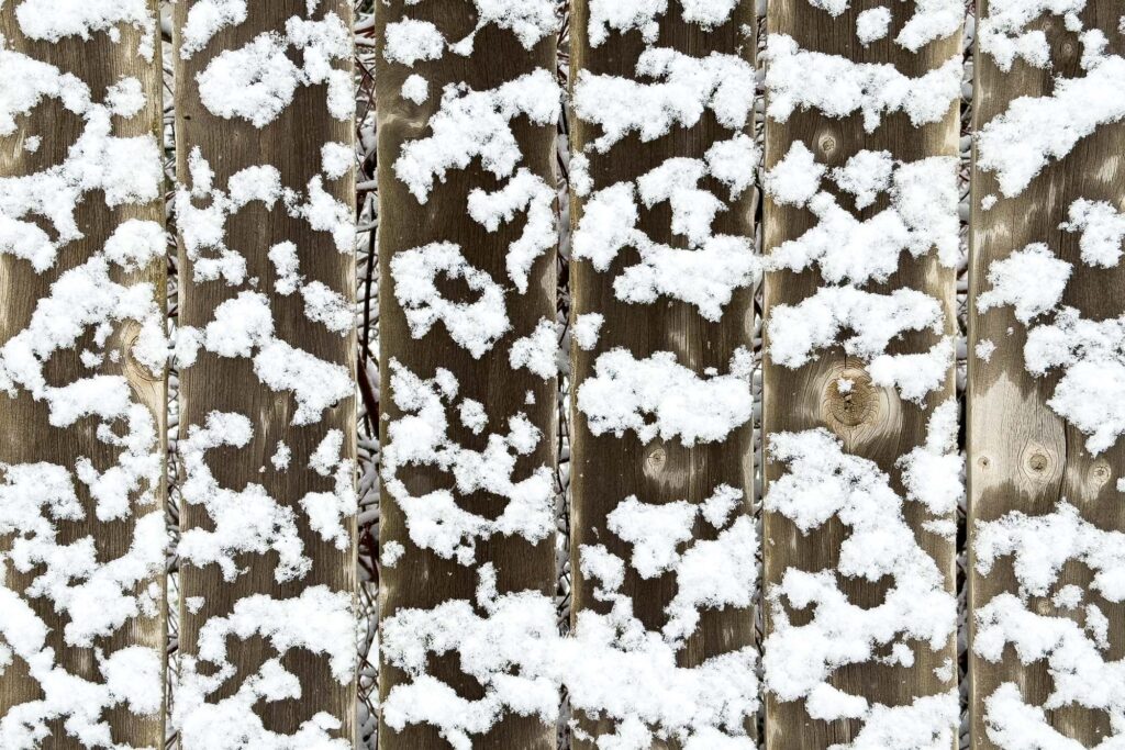Photograph of a weathered wooden fence with patches of fresh snow. Some of the wood is damp between the snow other areas it is dry and knotted.