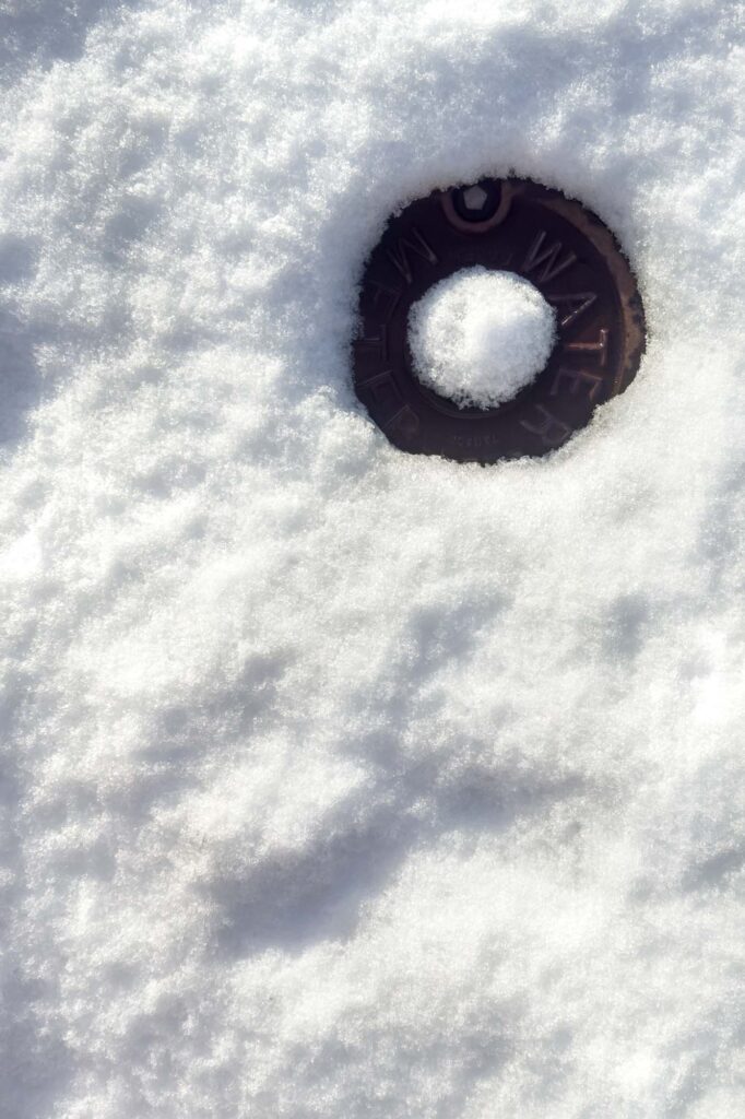Photograph of a circular water meter cover visible through lawn full of snow with a pile of snow in the middle of the water meter, like a moon showing through clouds.