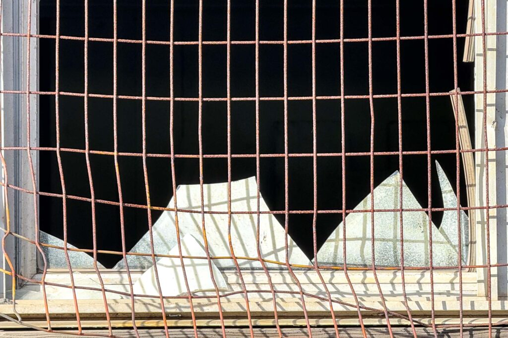 Photograph of rusted wire over a broken window with shards of glass covered in white paint.