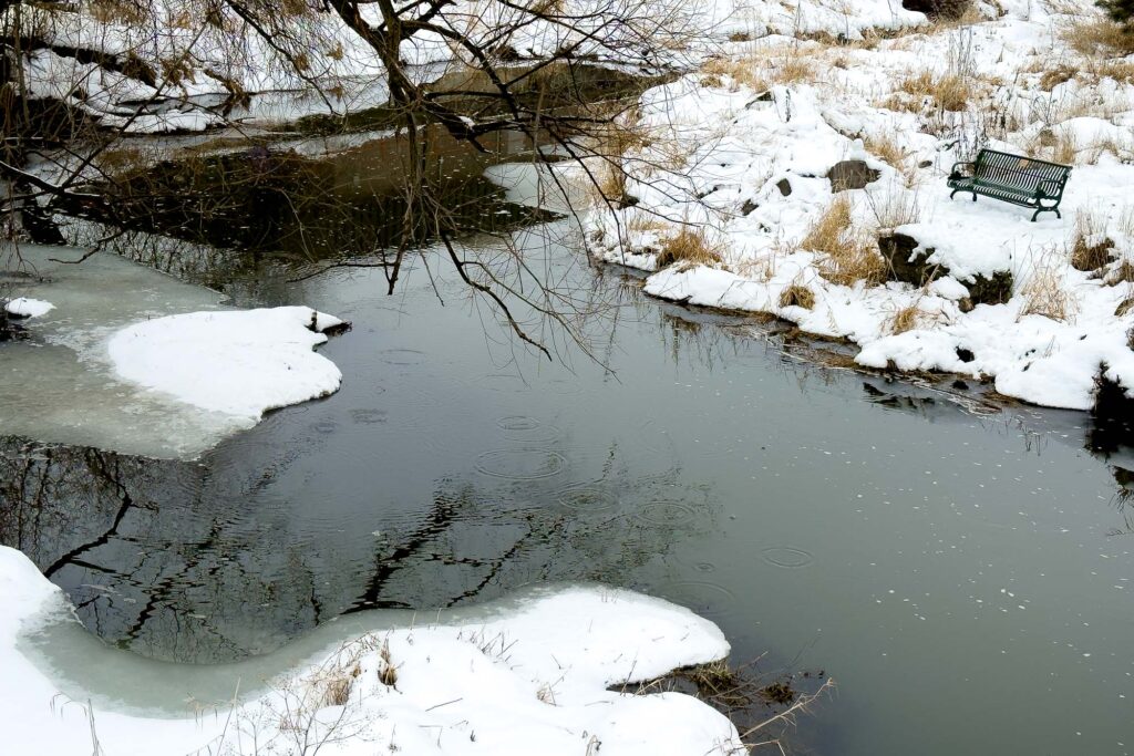 Photograph of a creek from a bridge with ripples from drops of melting snow from a tree, reflections of downtown buildings and an empty park bench on the shore.