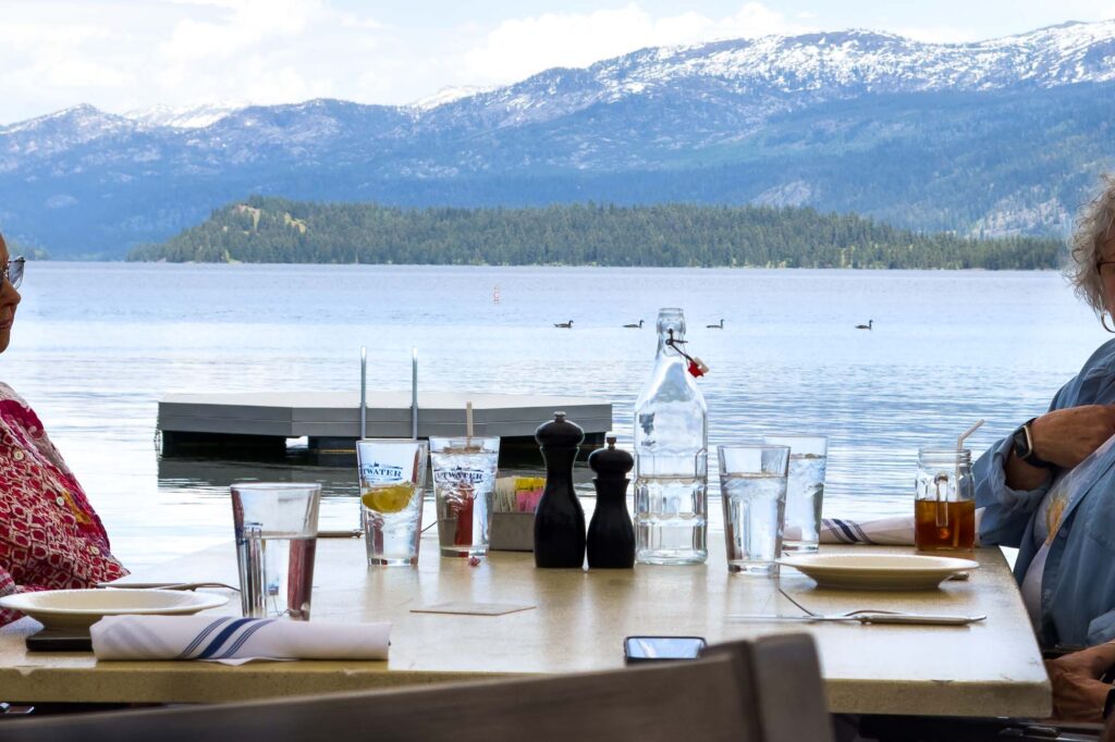 Color photograph of snow capped mountains above a lake, looking through an occupied table at a restaurant with glasses of clear water. Floating on the lake, geese pass by the top of a glass water bottle.
