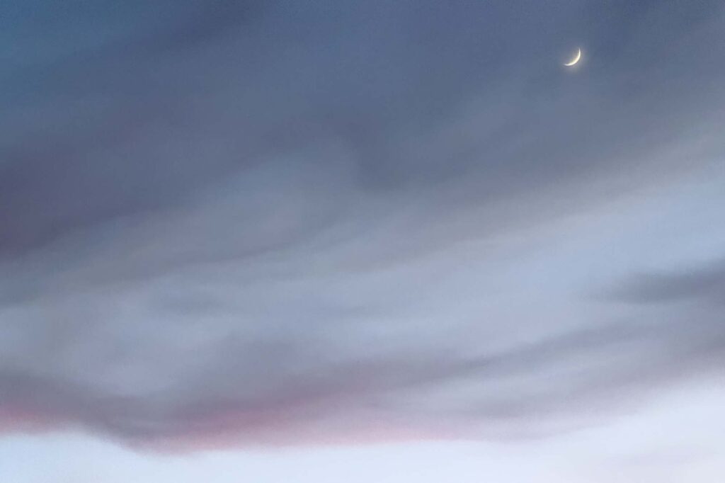 Photograph of the new moon and light evening clouds darkened at sunset, the lower clouds of magenta over a clear sky.