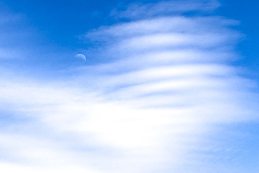 Color photograph of a new moon in a blue sky at the edge of a unique formation of lines, or like waves.