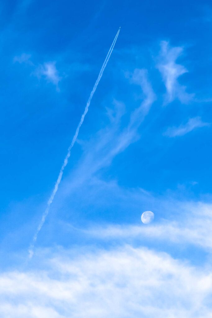 Color photograph of the moon in a blue sky surrounded by light clouds, a plane leaving a contrail is flies by.