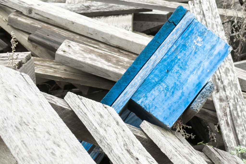Color photograph of a pile of wooden pallets, weathered, and grey, the edge of one pallet sticking out of the pile is painted blue.