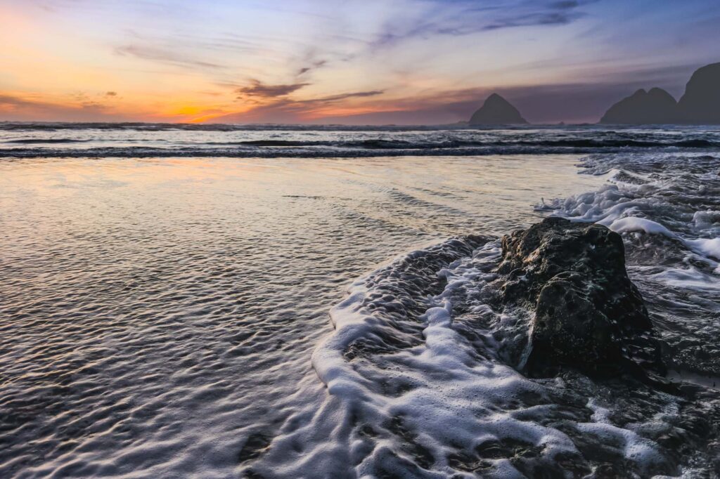 Color photograph of a sunset over the ocean with waves coming over a a  sandbar splashing into a rock.