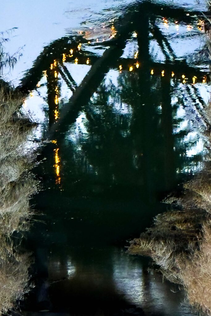 Color photograph of a creek reflecting a railroad bridge with a string of lights
