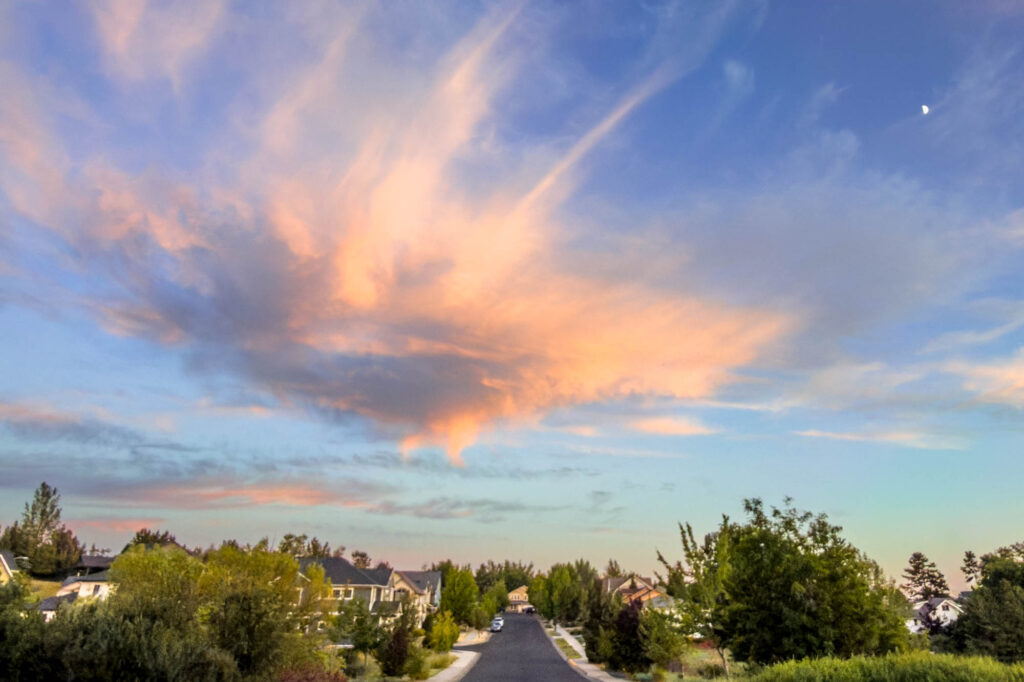 Color photograph of a unique shaped cloud at sunset, wispy shaped and orange in color, above an empty suburban street. A small quarter moon is high in the sky in the top right