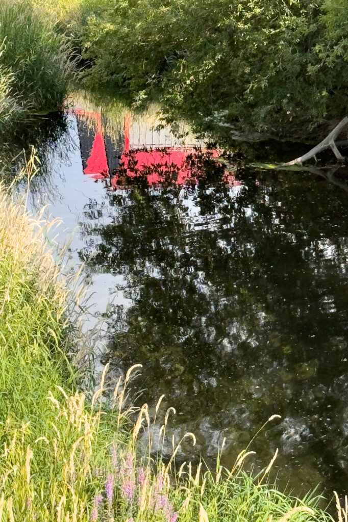 Color photograph of pooled water in a stream reflecting two barns, trees, and a blue sky. Trees, green grasses, and few flower blooms grow on the banks.
