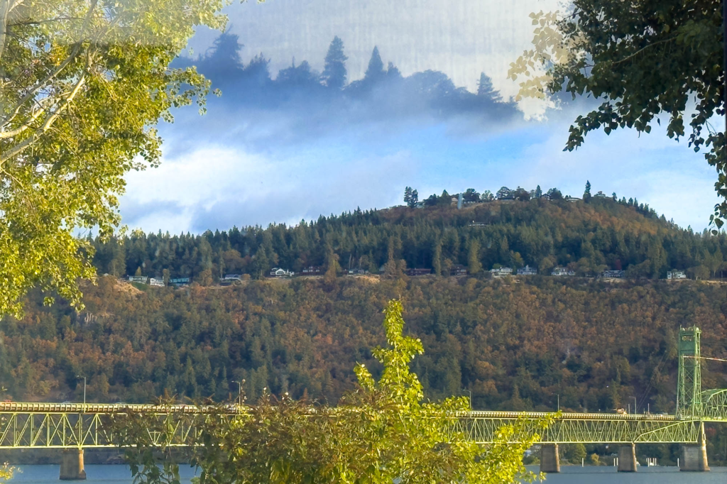 A color photograph of a river mountain scene, there is a green metal bridge going over the river, above it mountains and a blue sky with patchy dark clouds, above the blue sky and cloud is a mountain floating reflected in glass.